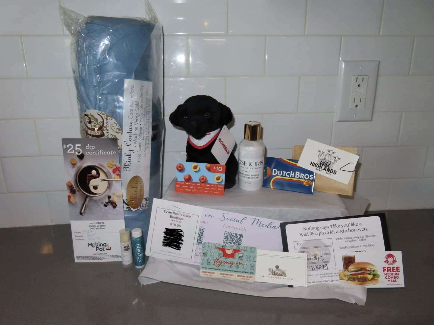 Items from a 2026 care basket laid out on a counter: a Minky Couture blanket, a plush dog, skincare, snacks, and gift cards from local businesses