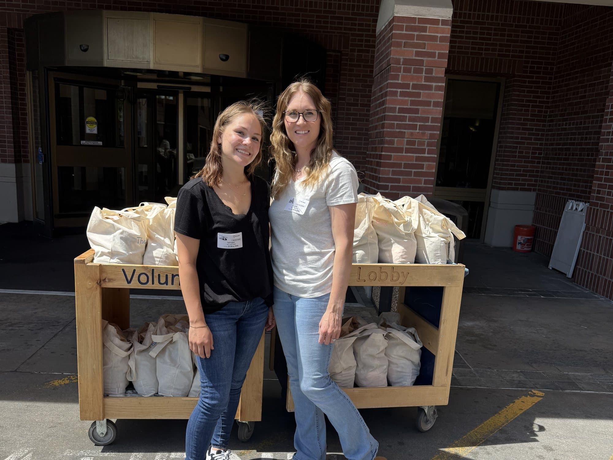 Tiffany and Alyssa Higbee standing in front of carts stacked with care-basket tote bags outside a hospital entrance