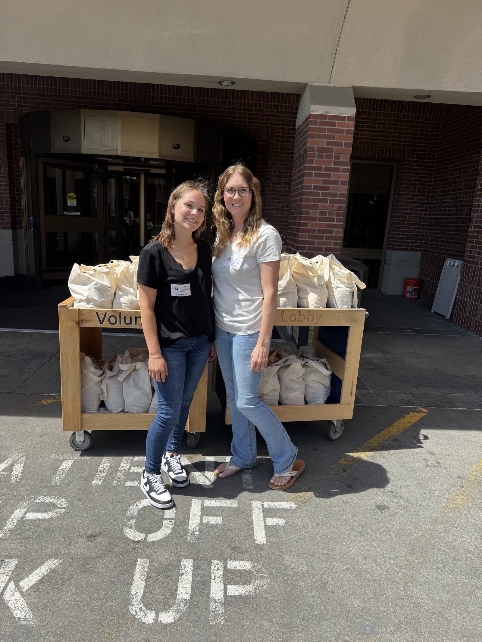 Tiffany and Alyssa Higbee standing in front of carts stacked with care-basket tote bags outside a hospital entrance