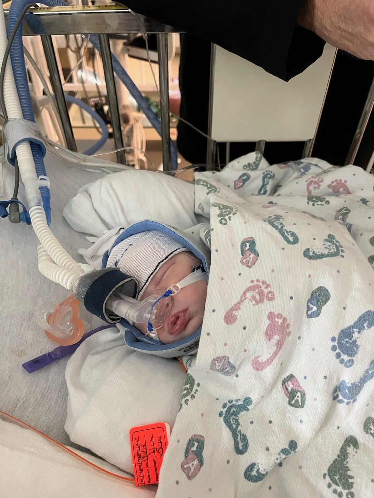 Maverick as a newborn in a hospital bed, wearing a knit hat and breathing support, under a footprint-print blanket